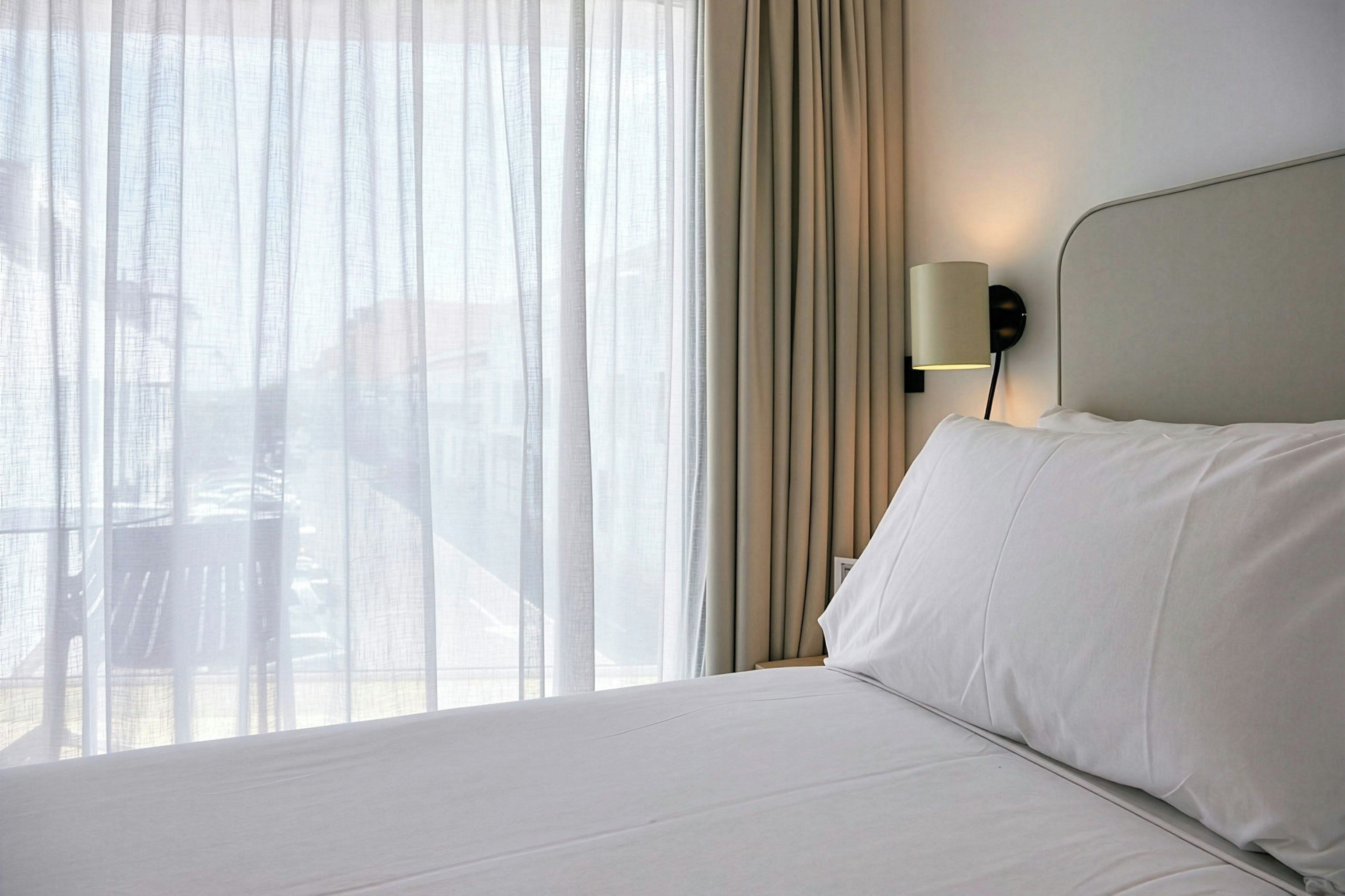 A hotel room with a bed, a nightstand, a lamp, and a view through sheer curtains.