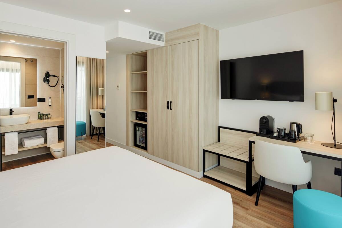 A modern hotel room with a large bed, a desk with a chair, a TV, and a wardrobe.