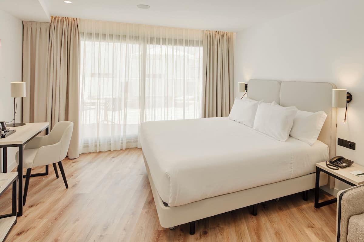 Una habitación de hotel con una cama grande, un escritorio, una silla y una mesita de noche con lámpara.