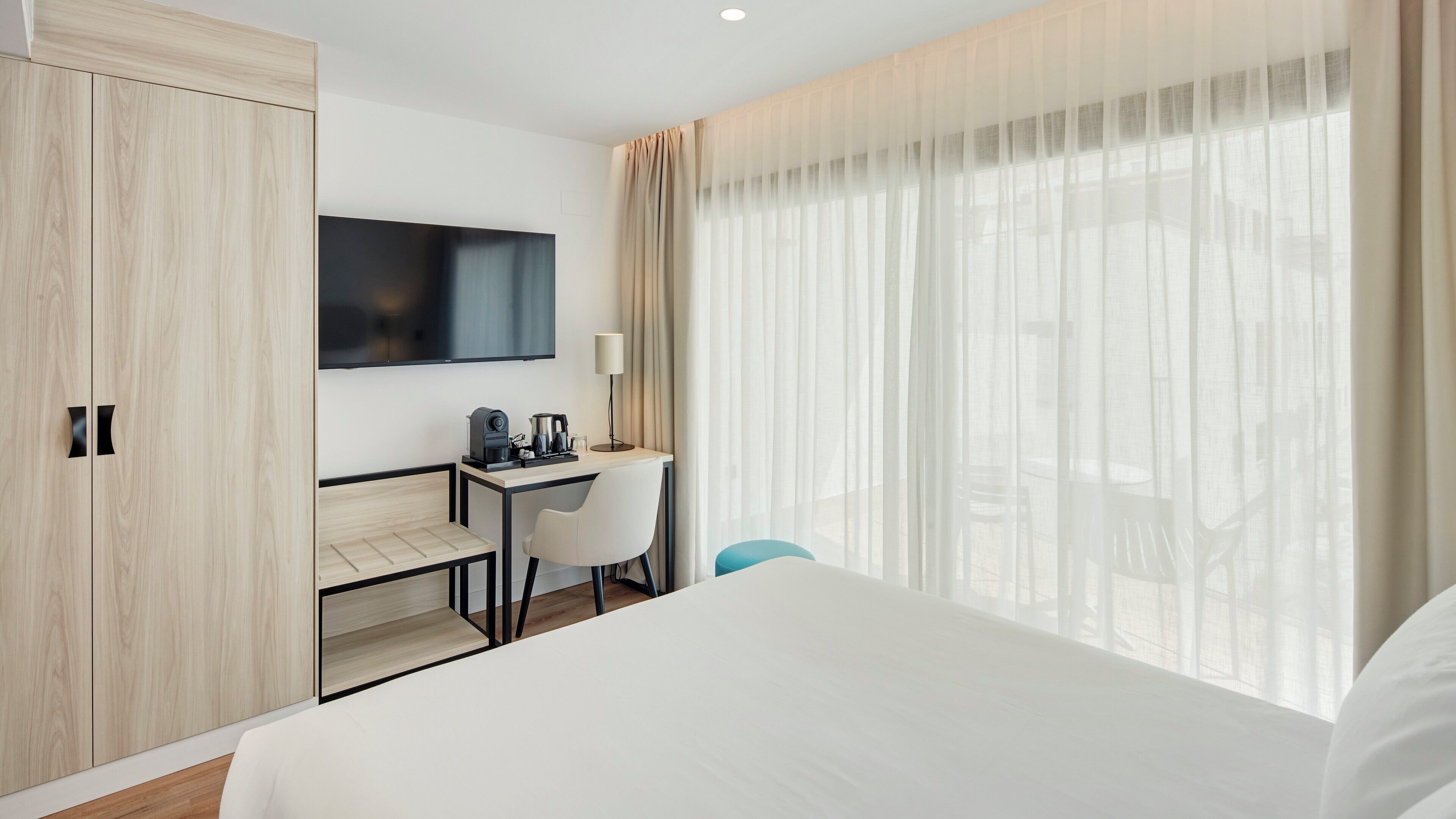 Habitación de hotel con cama, escritorio, silla, televisión y un amplio ventanal con cortinas.