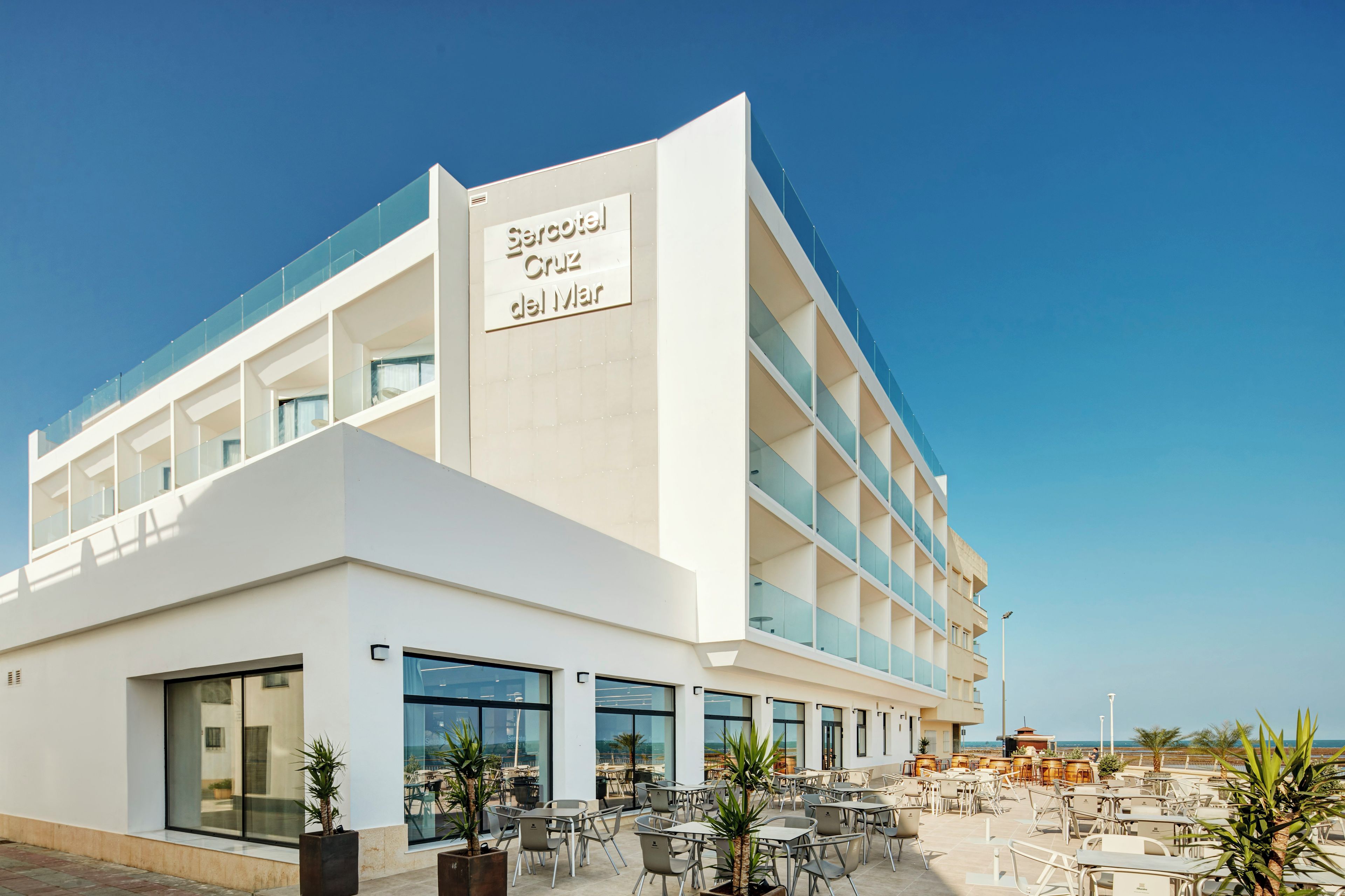 Un hotel moderno con un letrero llamativo que dice 'Hotel Cruz del Mar'. Cuenta con un amplio espacio al aire libre con mesas y sillas, y una vista despejada del mar al fondo.