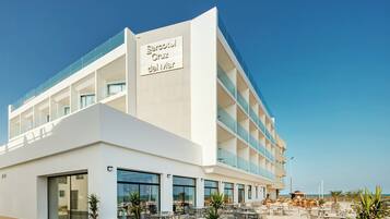 Un hotel moderno con un letrero llamativo que dice 'Hotel Cruz del Mar'. Cuenta con un amplio espacio al aire libre con mesas y sillas, y una vista despejada del mar al fondo.