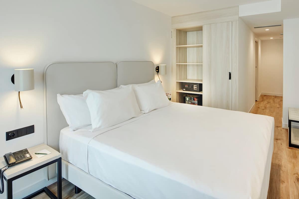 A modern hotel room with a large bed, bedside lamps, a desk with a telephone, and a wardrobe with a mirror.