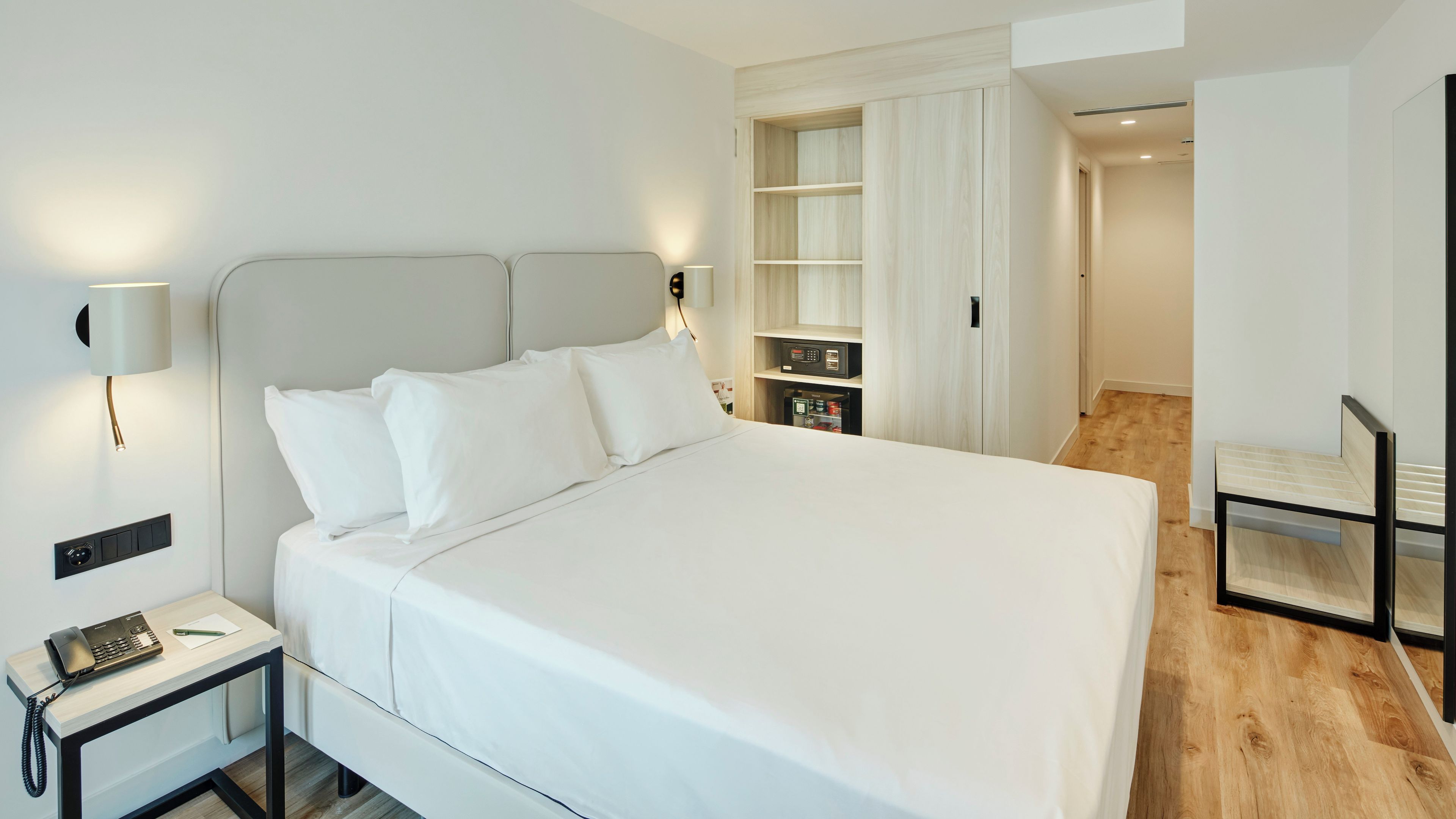 A neatly made bed with white linens, a bedside table with a telephone, and a wall-mounted lamp.