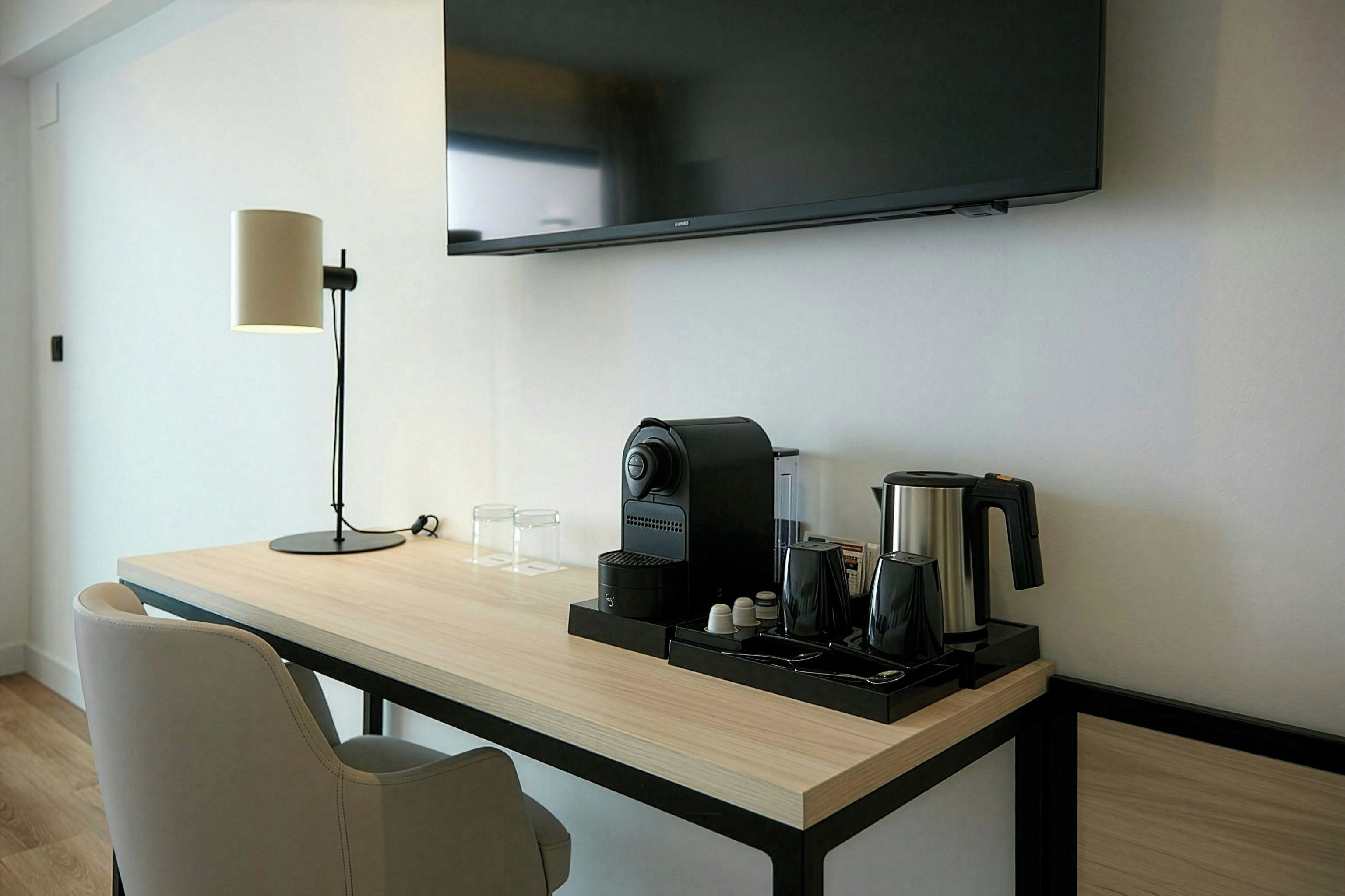 A hotel room with a wooden desk, a coffee maker, a kettle, a lamp, and a flat-screen TV mounted on the wall.
