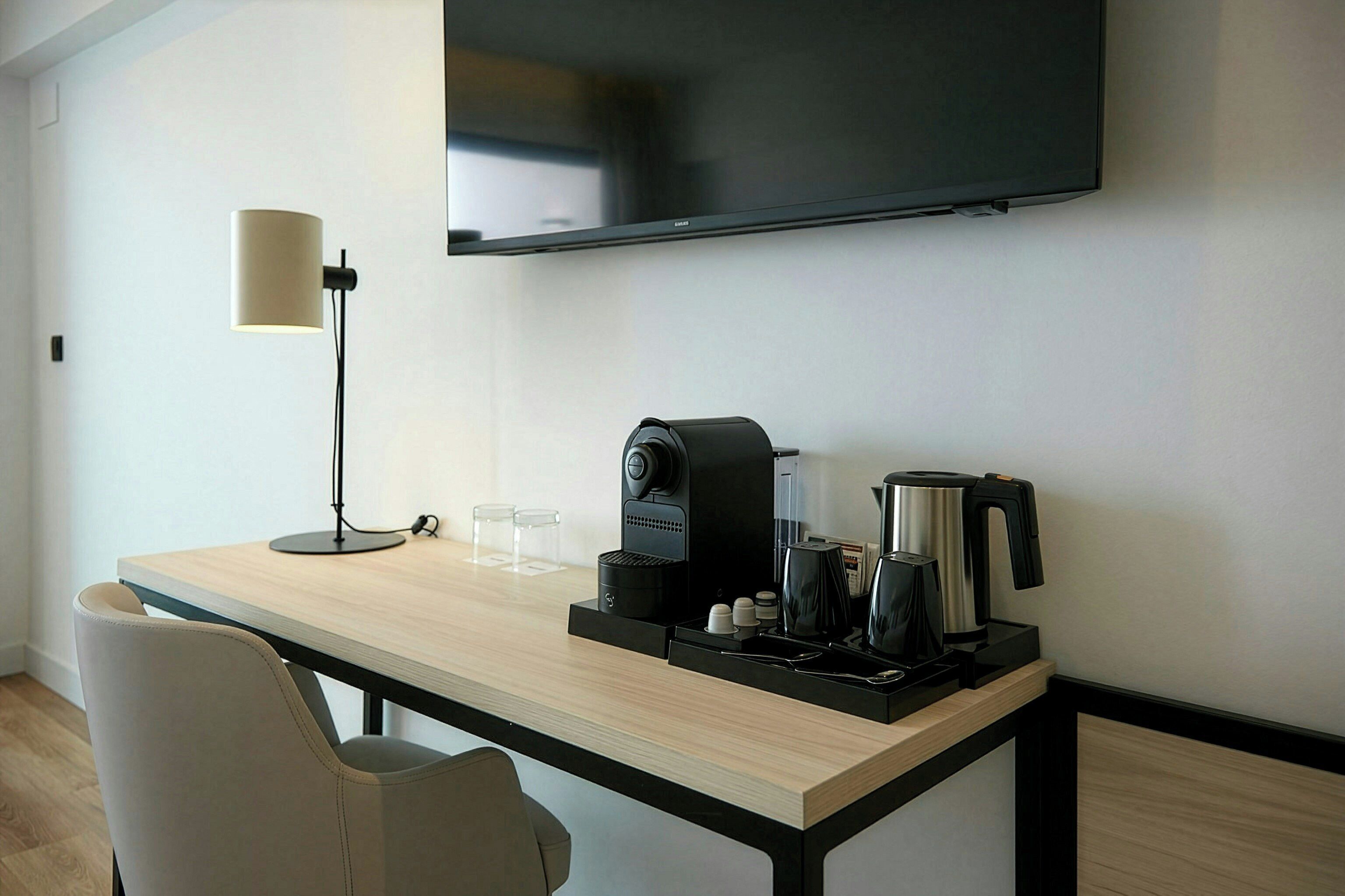 A hotel room with a wooden desk, a coffee maker, a kettle, a flat-screen TV mounted on the wall, a floor lamp, and a chair.