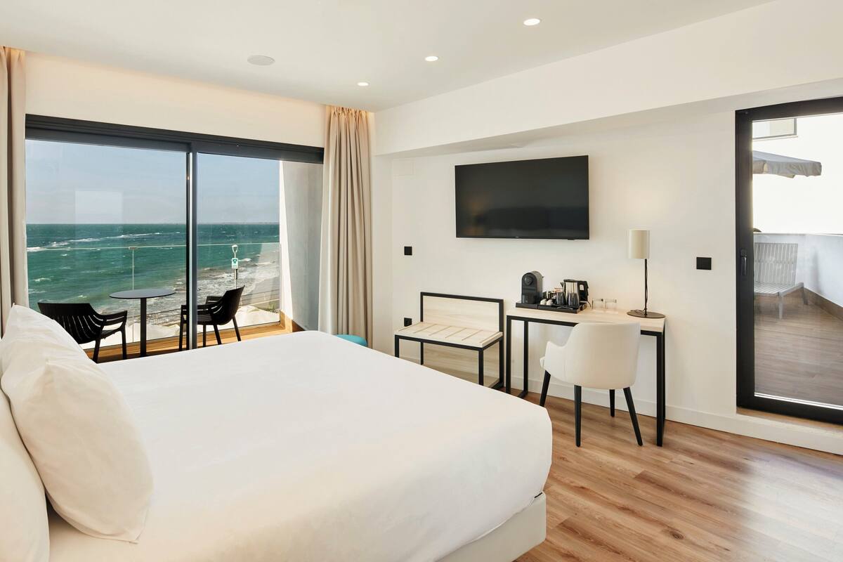 A hotel room with a large bed, a desk with a chair, a TV, and a view of the ocean.