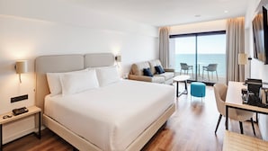 A hotel room with a large bed, a desk with a chair, a small table, and a sofa. There is a large window with a view of the sea. - Sercotel Cruz del Mar (Chipiona)