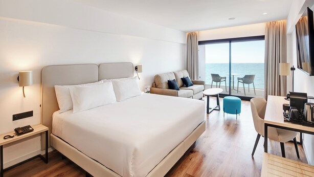 A hotel room with a large bed, a desk with a chair, a small table, and a sofa. There is a large window with a view of the sea. - Sercotel Cruz del Mar  (Chipiona)