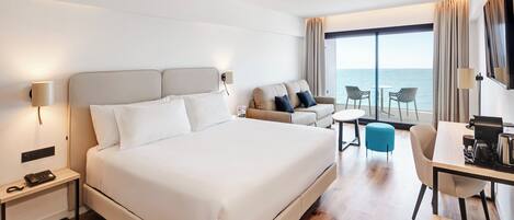 Ein Hotelzimmer mit einem großen Bett, einem Schreibtisch mit Stuhl, einem kleinen Tisch und einer Couch. Ein großes Fenster bietet Blick auf das Meer.