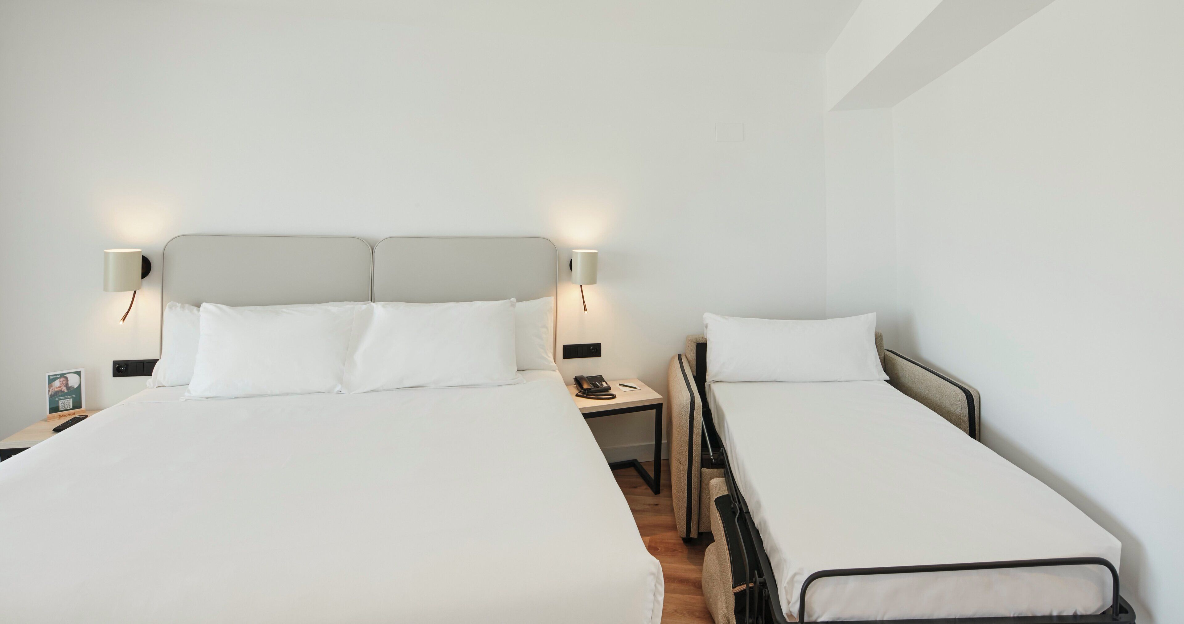 A hotel room with two beds, each with white linens and headboards. There are bedside tables with lamps and a phone.