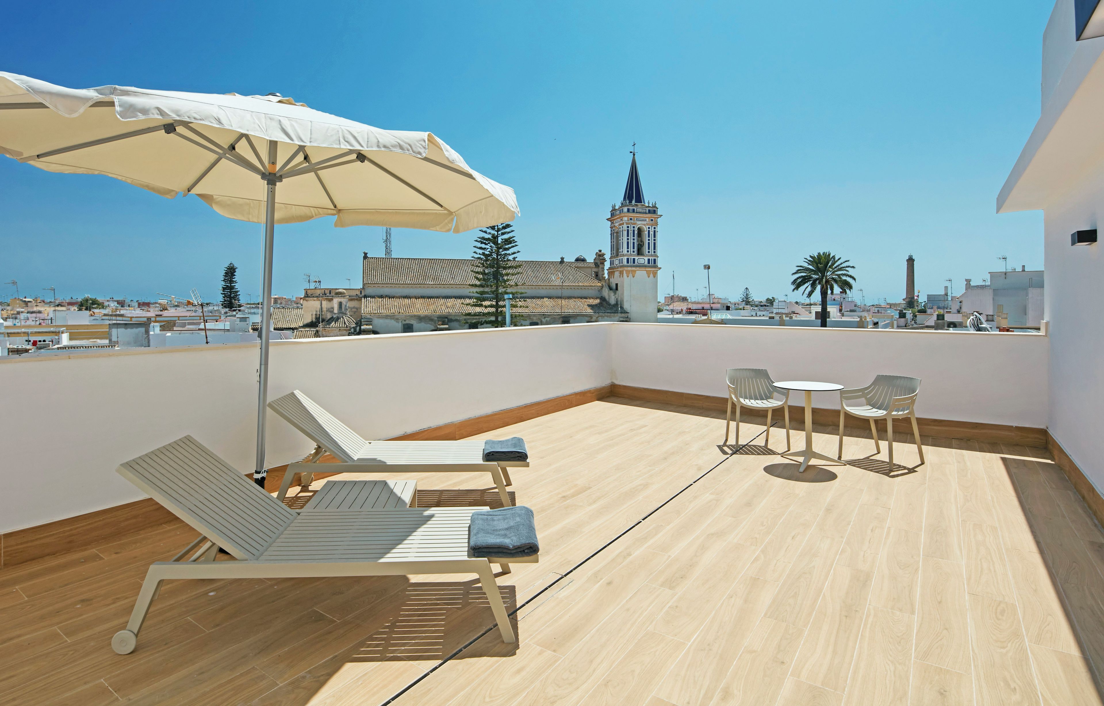 Una terraza en la azotea con una sombrilla blanca, sillones de descanso y una mesa con sillas. Se observa un edificio histórico con una torre al fondo.