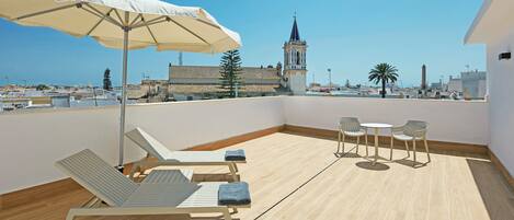A rooftop terrace with a white umbrella, lounge chairs, and a table with chairs. A historic building with a tower is visible in the background.