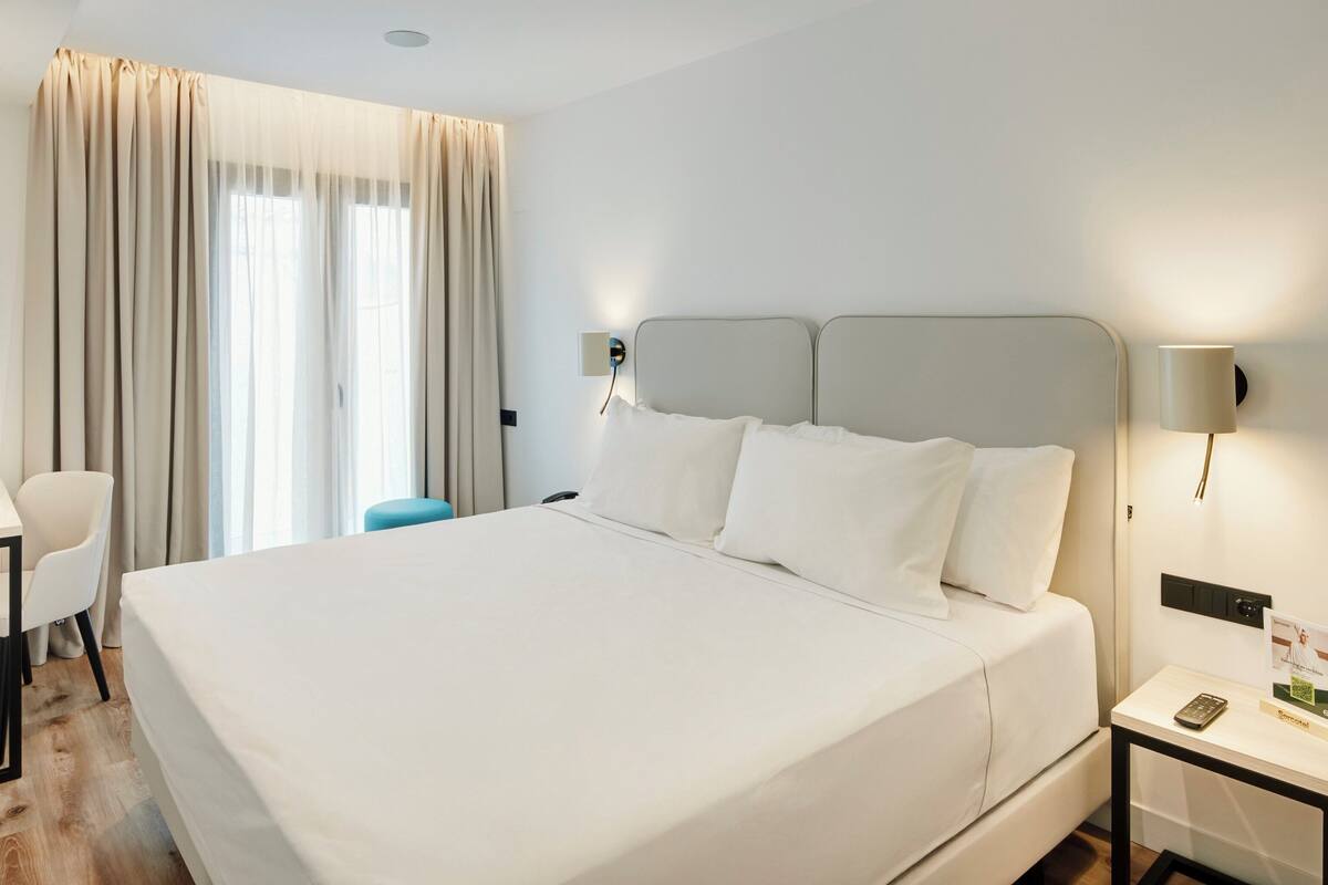 A hotel room with a large bed, a desk with a chair, a small table, and a nightstand with a lamp.