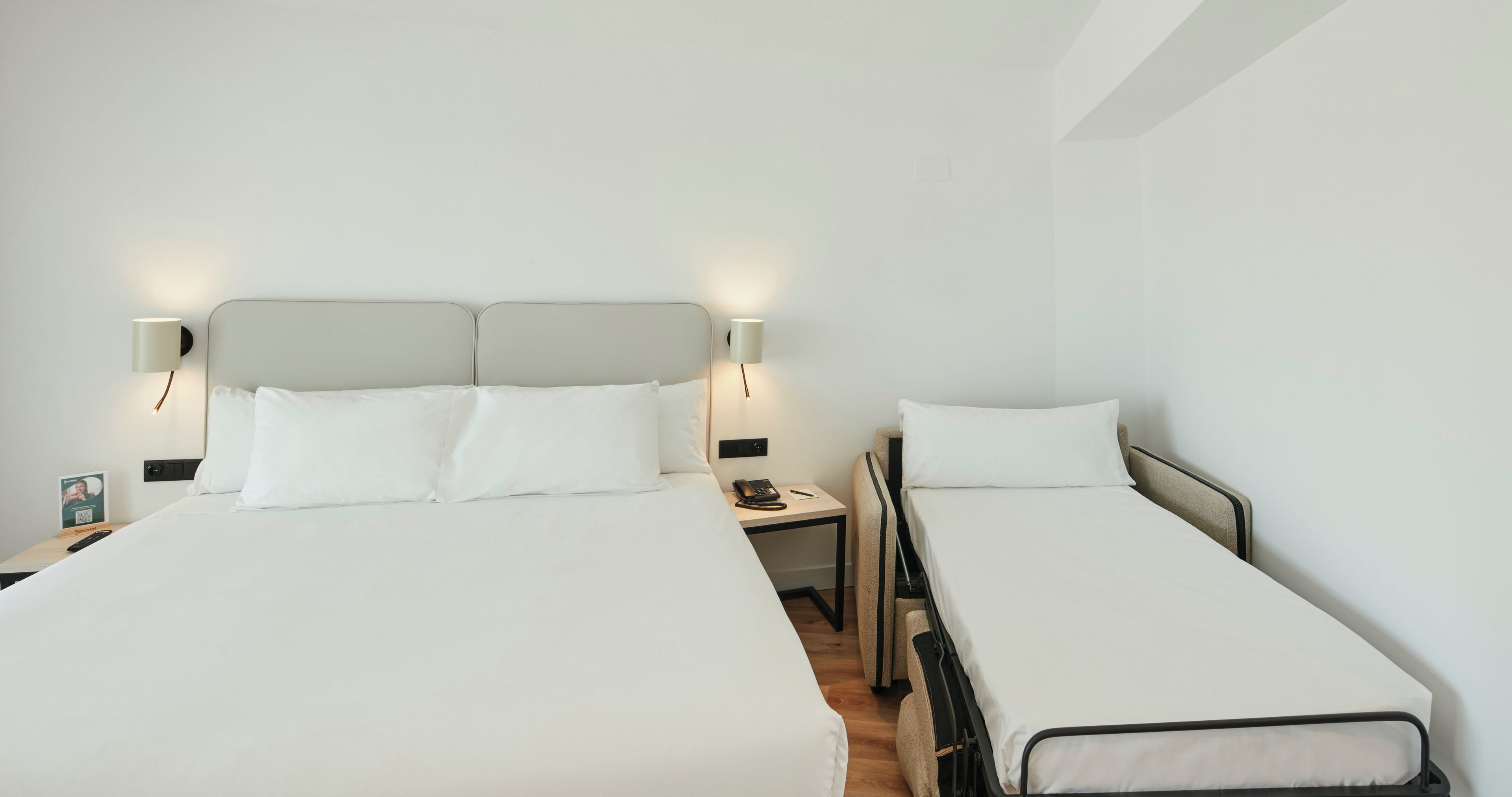 Habitación de hotel con dos camas, cada una con ropa blanca y cabeceros. Se observan mesitas de noche con lámparas y un teléfono.
