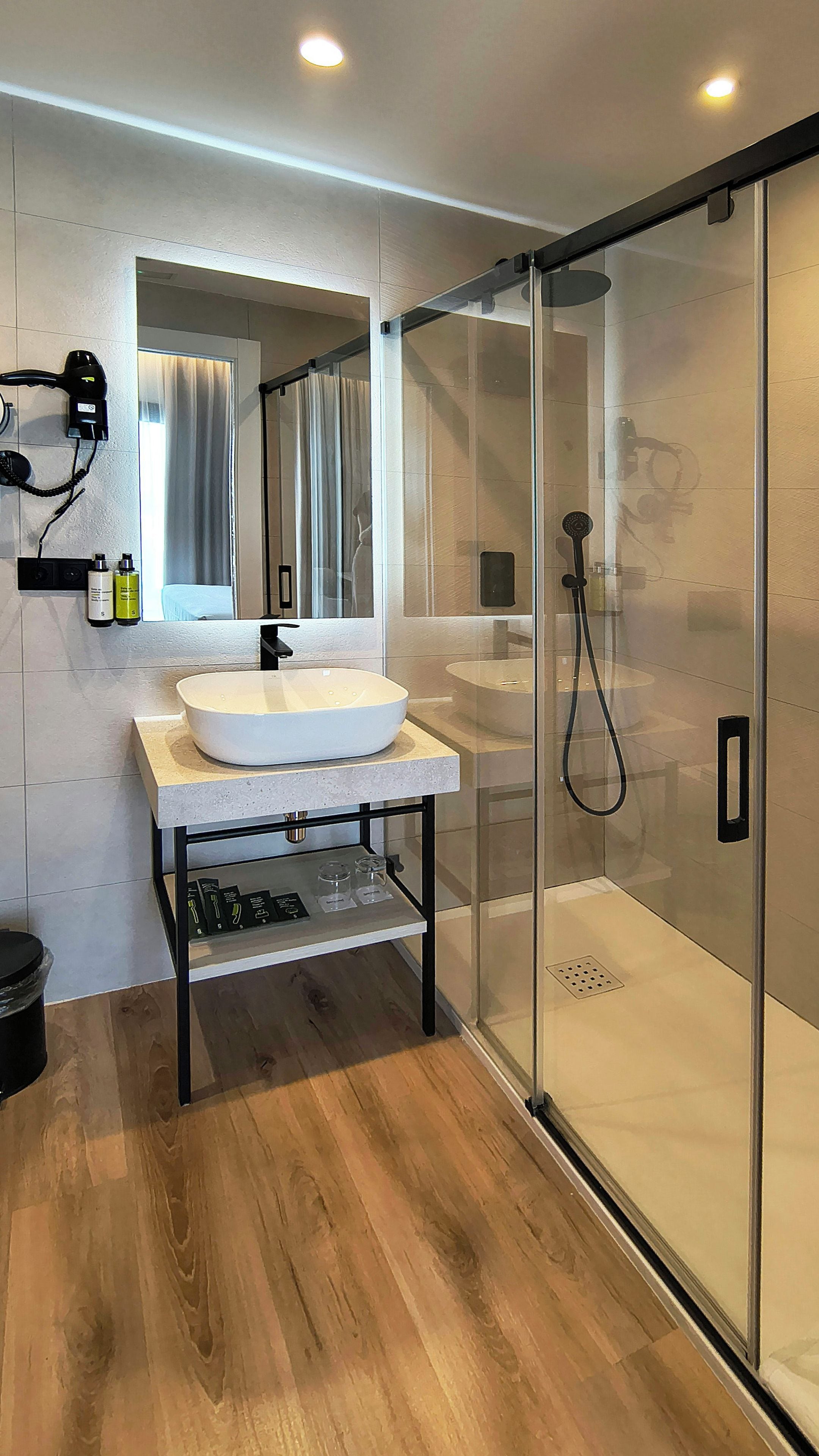 Un baño moderno con ducha envuelta en vidrio, lavamanos con espejo y secador de pelo montado en la pared.