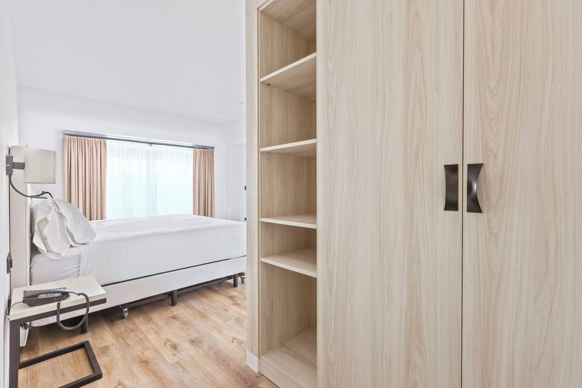 A hotel room with a bed, a bedside table with a lamp, a wardrobe, and a window with curtains.