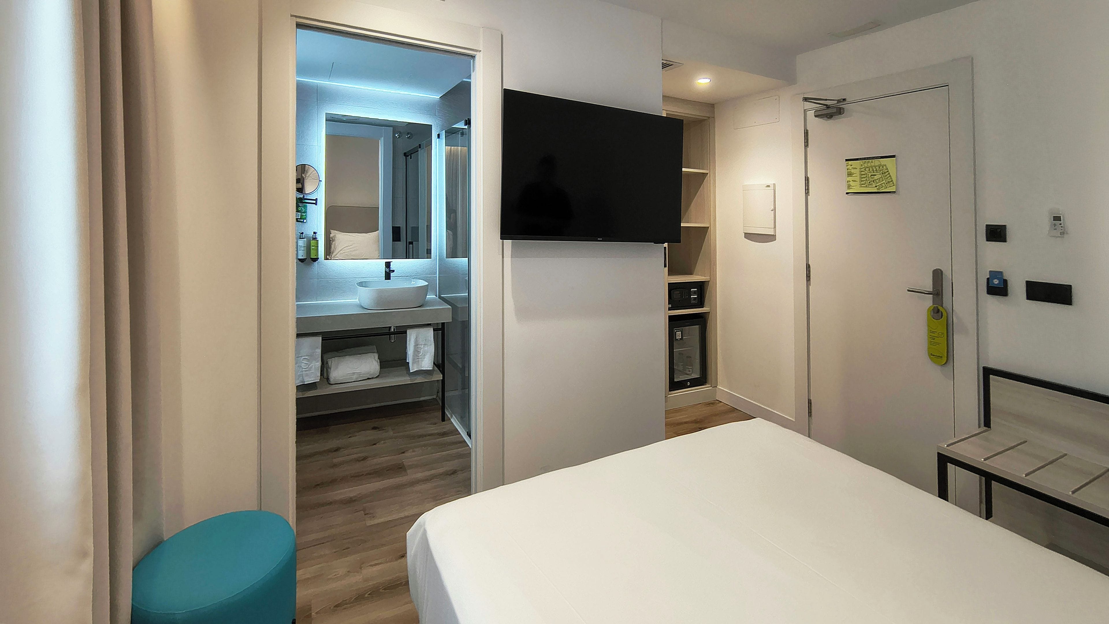 A hotel room with a bed, a TV mounted on the wall, a bathroom with a sink and mirror, and a door with a lock.