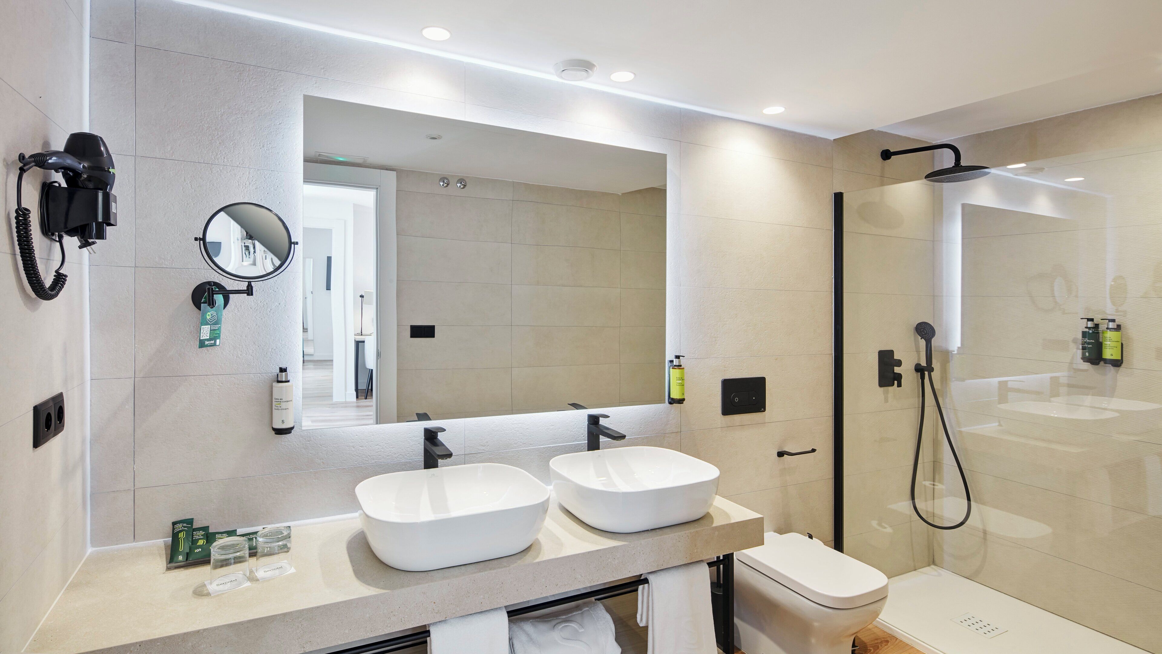 Un baño moderno con dos lavamanos, un espejo grande, un cabezal de ducha manual y un inodoro.