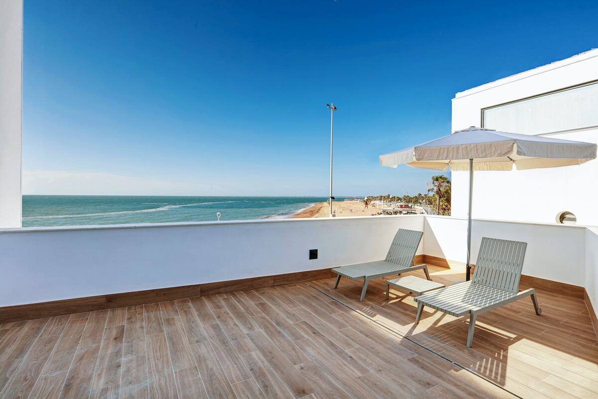 A balcony with a view of the beach, two chairs, and a white umbrella.