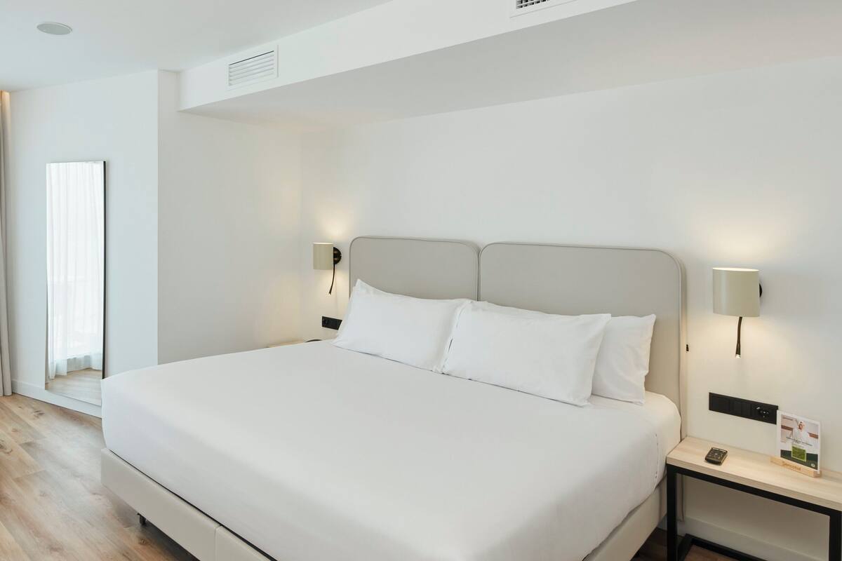 A modern hotel room with a large bed, two bedside tables, a nightstand, a small blue chair, and a large window with sheer curtains.