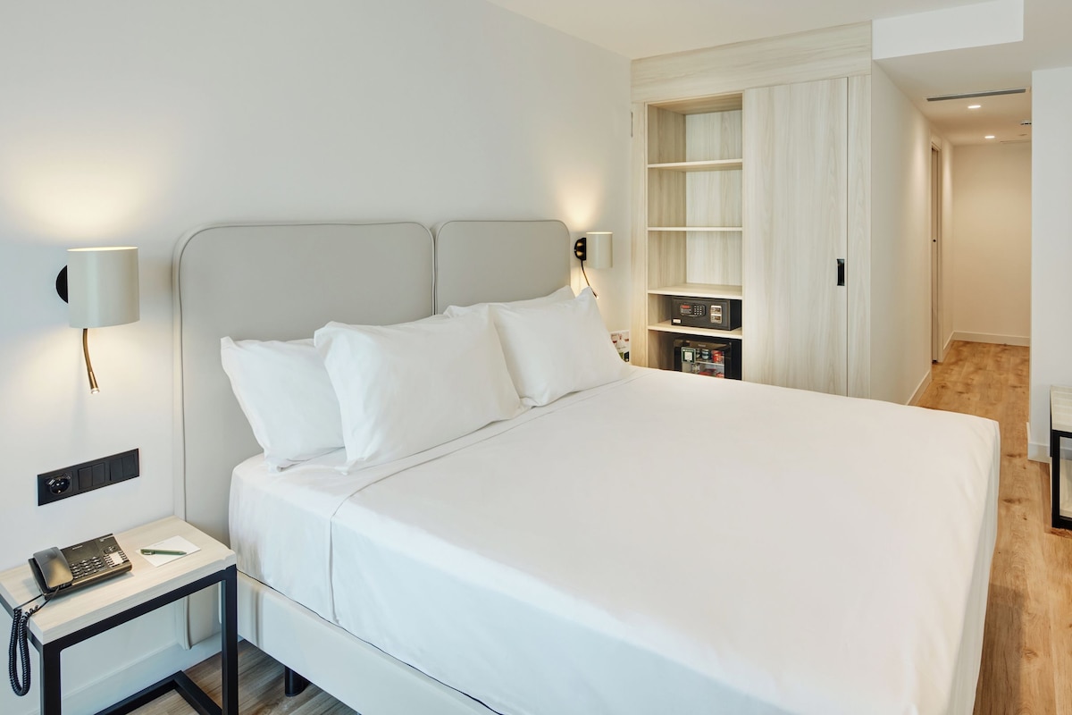 A neatly made bed with white linens, a bedside table with a telephone and a pen, and a wall-mounted lamp.