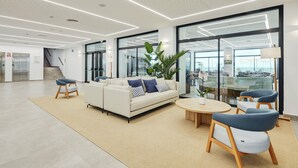 A modern lobby with a white sofa, blue chairs, a round wooden table, and large windows offering a view of the outdoors. - Sercotel Cruz del Mar (Chipiona)
