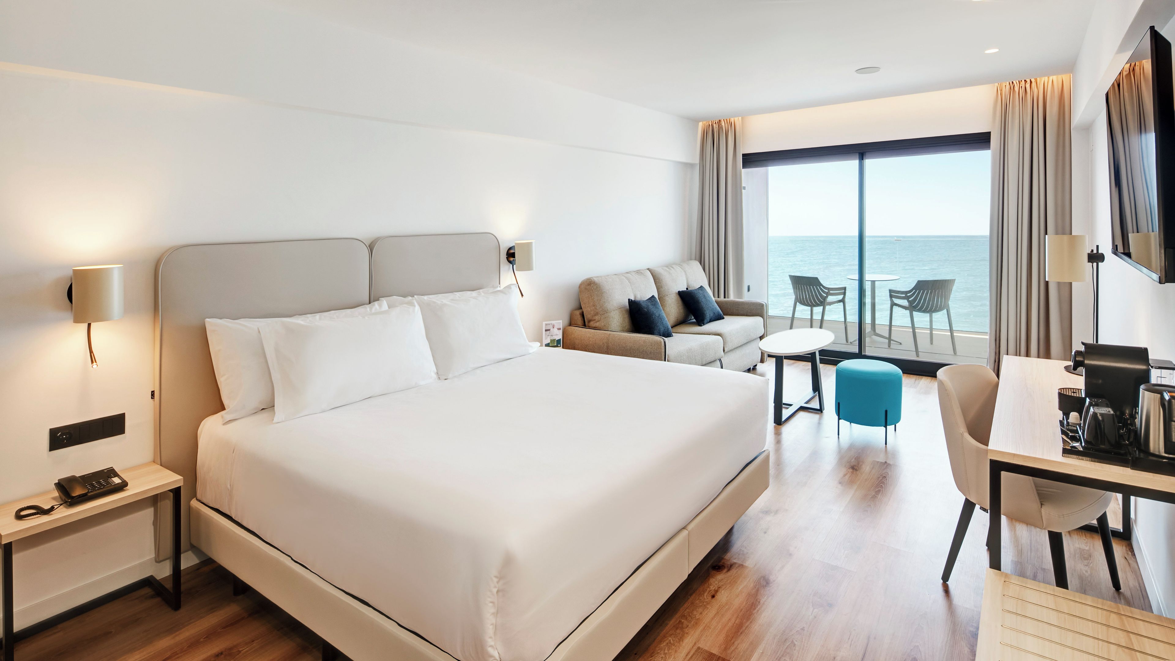 Een hotelkamer met een groot bed, een bureau, een stoel, een klein tafeltje en uitzicht op de zee door grote ramen.