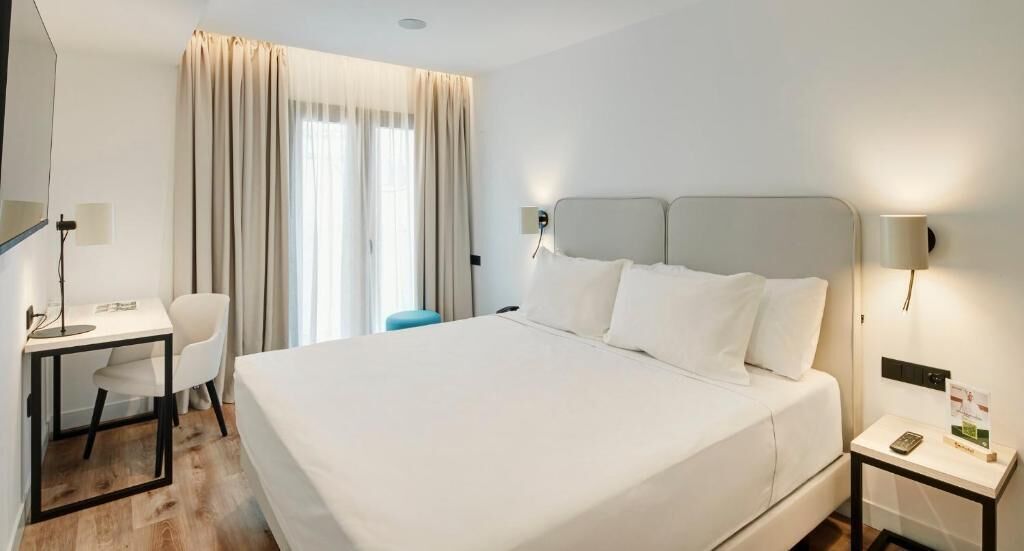 A hotel room with a large bed, a desk, a chair, a nightstand, and a window with curtains.