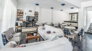 A modern living room with a fireplace, seating area, and a kitchen in the background. - DELLIS (Kamena Vourla)