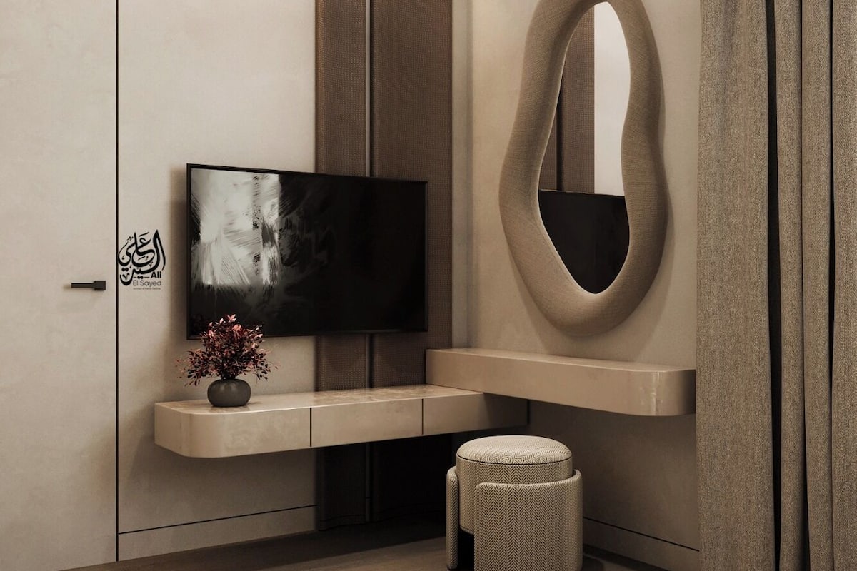 A modern bedroom with a flat-screen TV, a large mirror, a vanity, and a small ottoman.