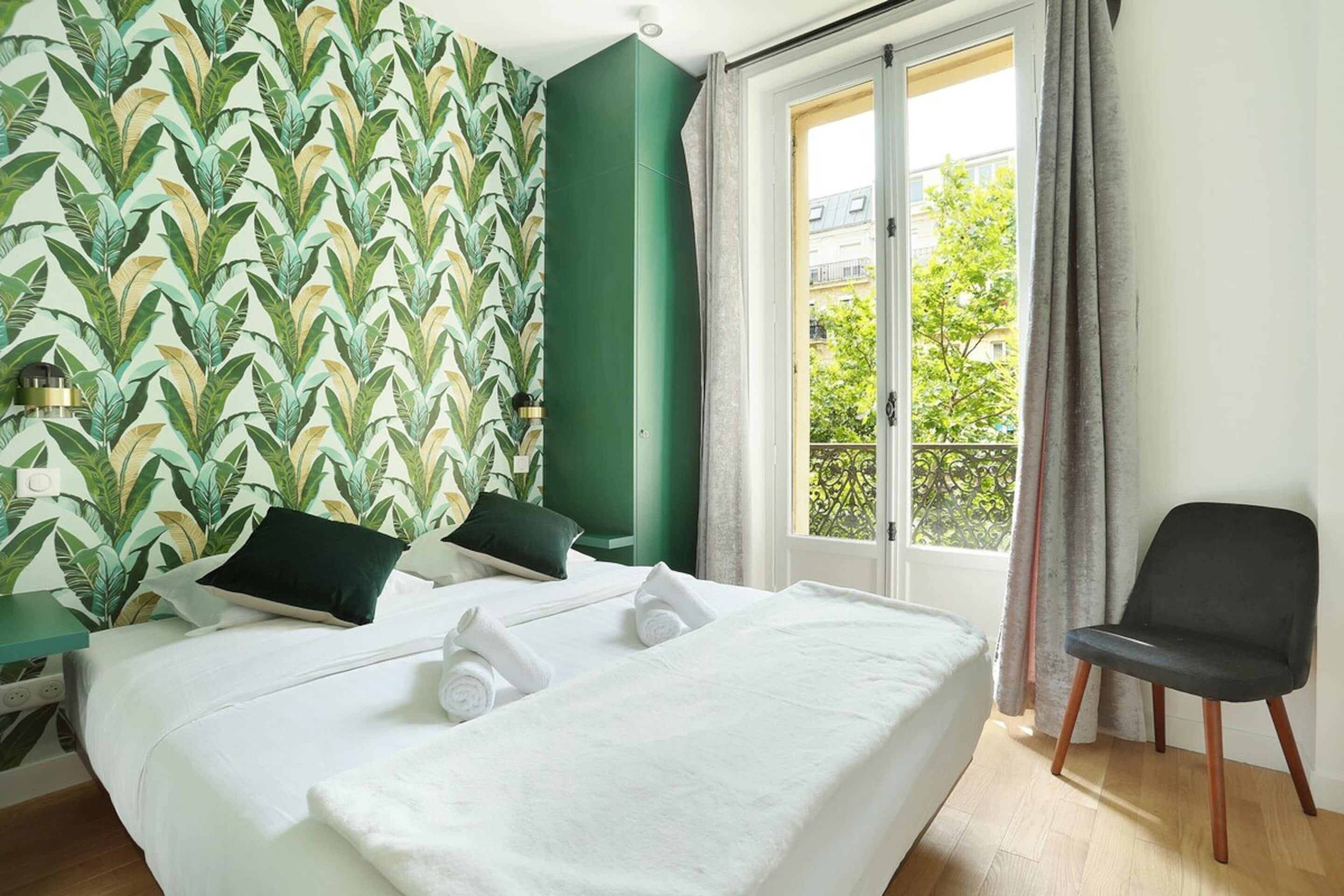 A hotel room with a bed, a chair, a window with curtains, and a green wall with a leaf pattern.