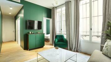 A modern living room with a green accent wall, a flat-screen TV mounted above a dark green cabinet, a green armchair, and a beige sofa with a decorative pillow.