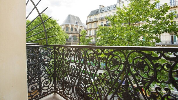 Ein Balkon mit aufwendiger schmiedeeiserner Geländer, der auf eine Straße mit historischen Gebäuden und üppigem Grün blickt.