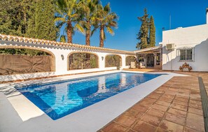 A swimming pool with a tiled deck, surrounded by a white building with arched windows and a covered patio area. - Awesome home in Guaro (Guaro)