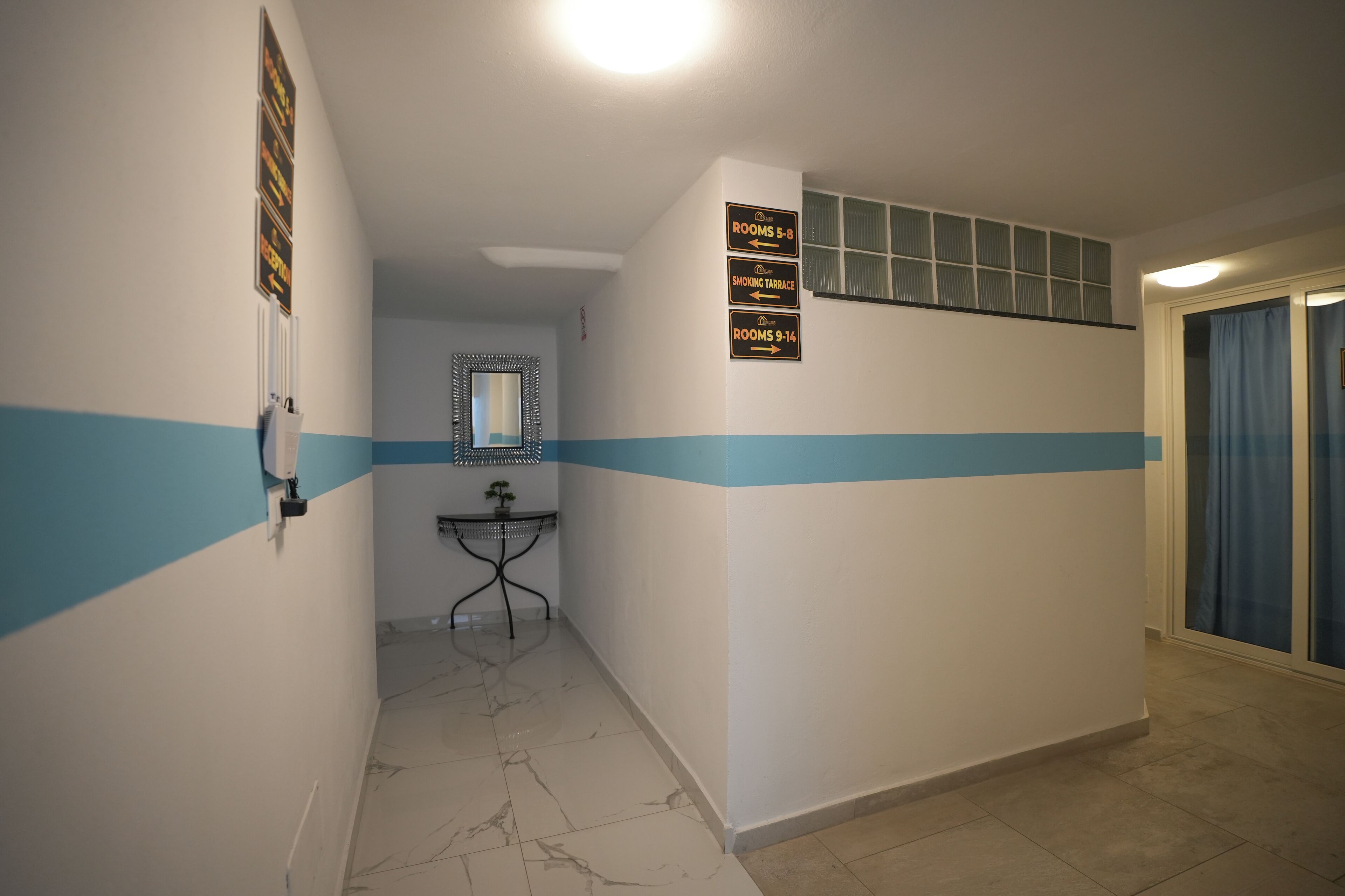 Corredor com placas indicativas para os quartos 5-9 e 9-14, um espelho e uma pequena mesa com uma planta.