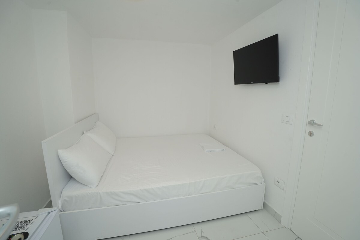 A small, white, single-bed room with a flat-screen TV mounted on the wall, a door, and a small table with a lamp.