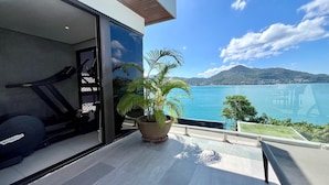 Une chambre moderne équipée d’un tapis de course, d’une grande plante en pot et d’un balcon donnant sur un paysage magnifique, avec un plan d’eau et des montagnes en toile de fond.
