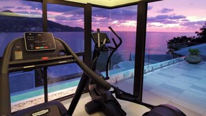 Une salle de sport à domicile équipée d’un tapis de course et d’un vélo elliptique, de grandes fenêtres offrant une vue sur le coucher de soleil sur l’eau, et d’une petite piscine intérieure.