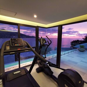 Een huisgymnasium met een loopband en een elliptische machine, grote ramen met uitzicht op een zonsondergang boven het water, en een klein binnenzwembad.