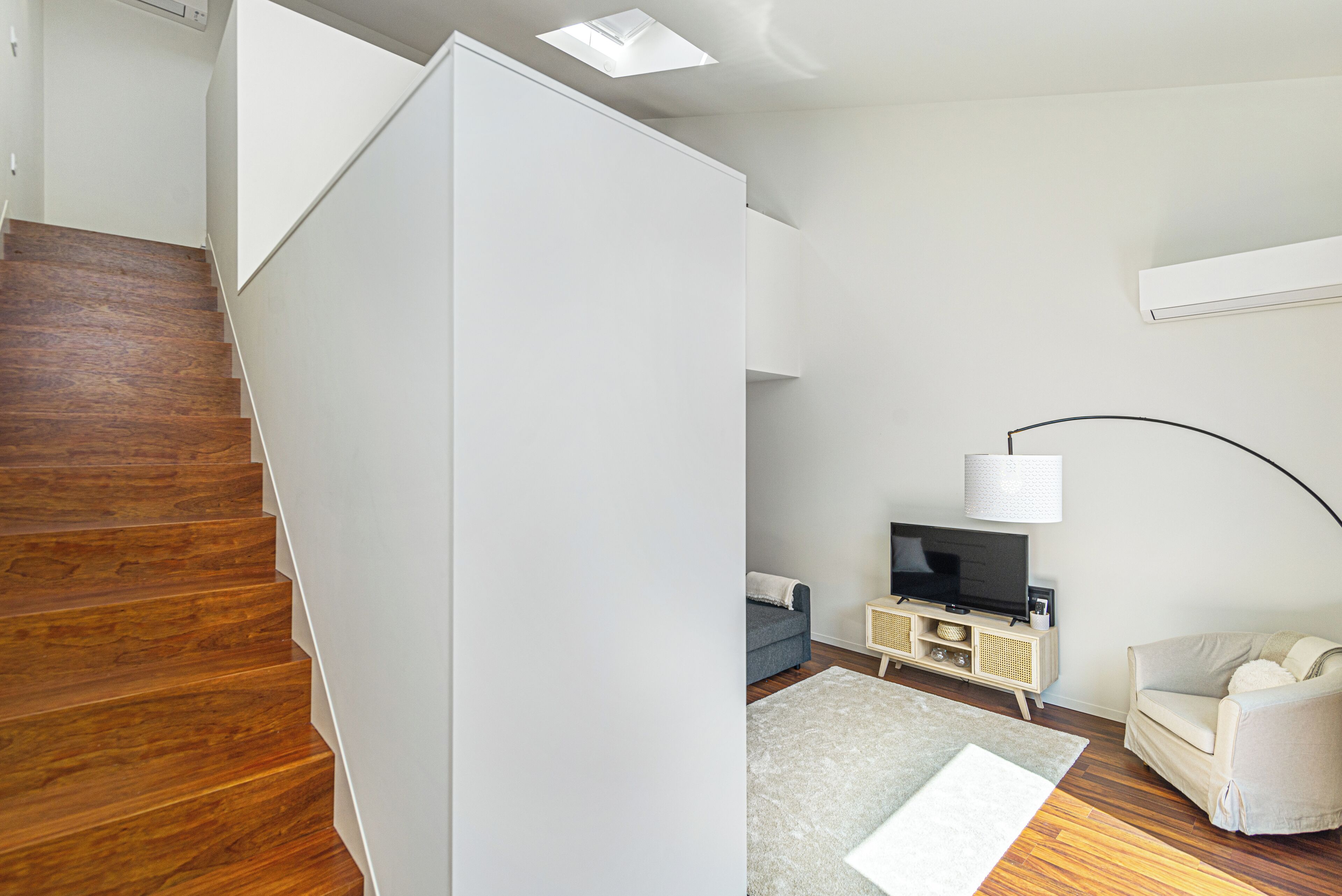 A modern living room with a wooden staircase, a flat-screen TV, a sofa, and a lamp.