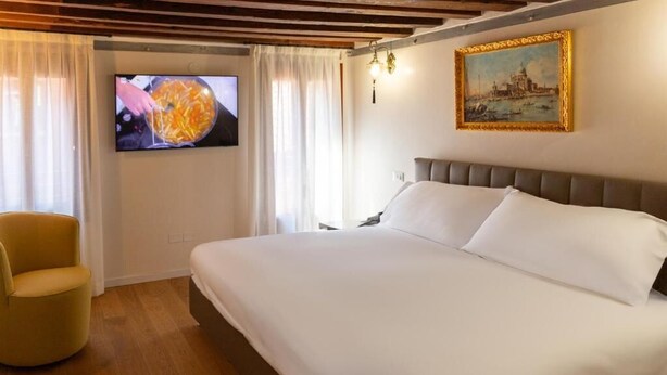Quarto de hotel com uma cama grande, uma televisão exibindo comida, uma cadeira e um quadro na parede.