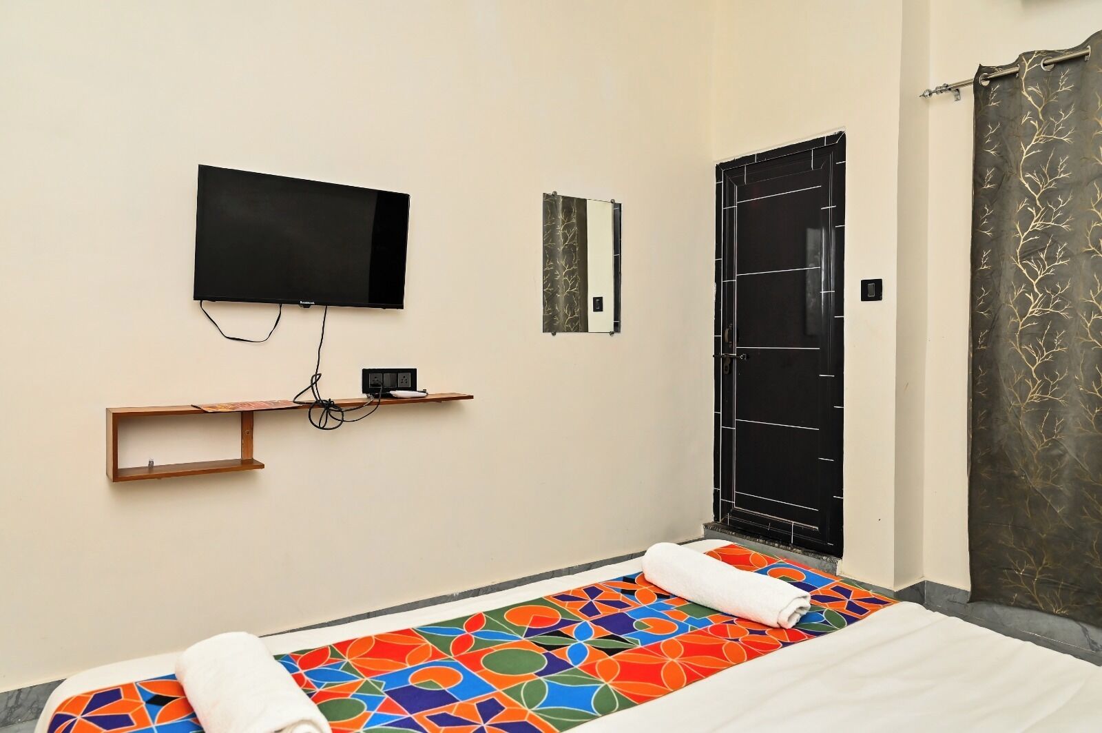 Una habitación con una cama, un televisor en la pared, una puerta con marco negro y una alfombra estampada.