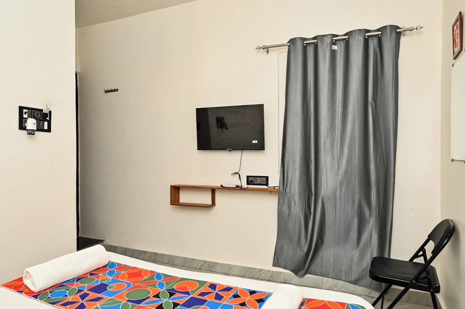 A bedroom with a bed, a TV mounted on the wall, a curtain, a chair, and a shelf with a remote control.