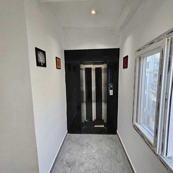 A hallway with a marble floor, an elevator, and a window with blinds. - The Shivaay Palace (Varanasi)