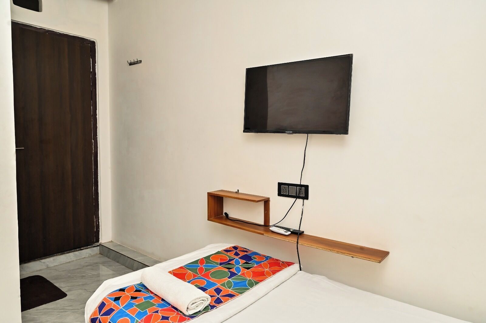 A room with a bed, a TV mounted on the wall, a wooden shelf, and a door.