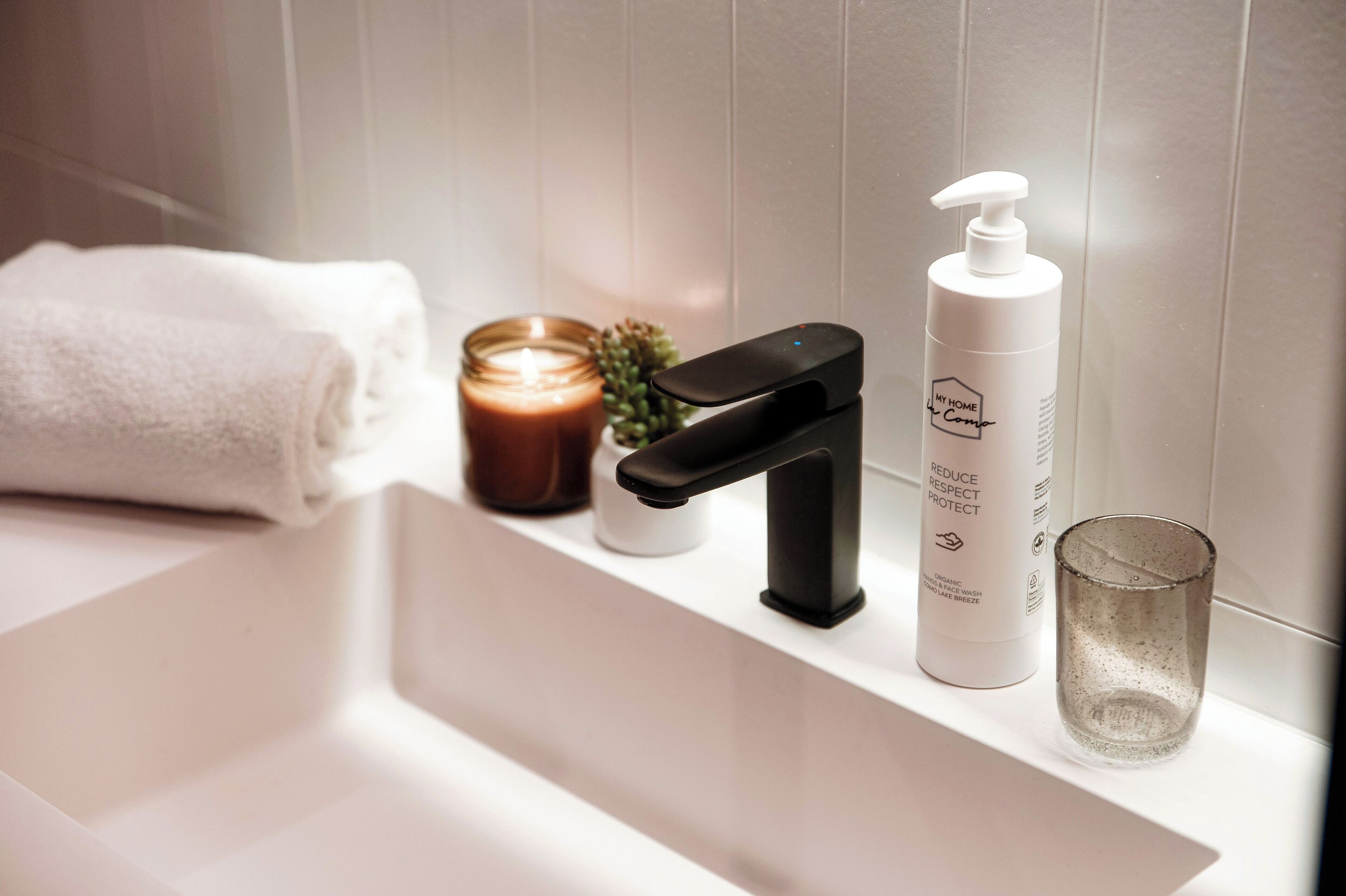 A bathroom sink with a black faucet, a white bottle of soap, a lit candle, and a clear glass.