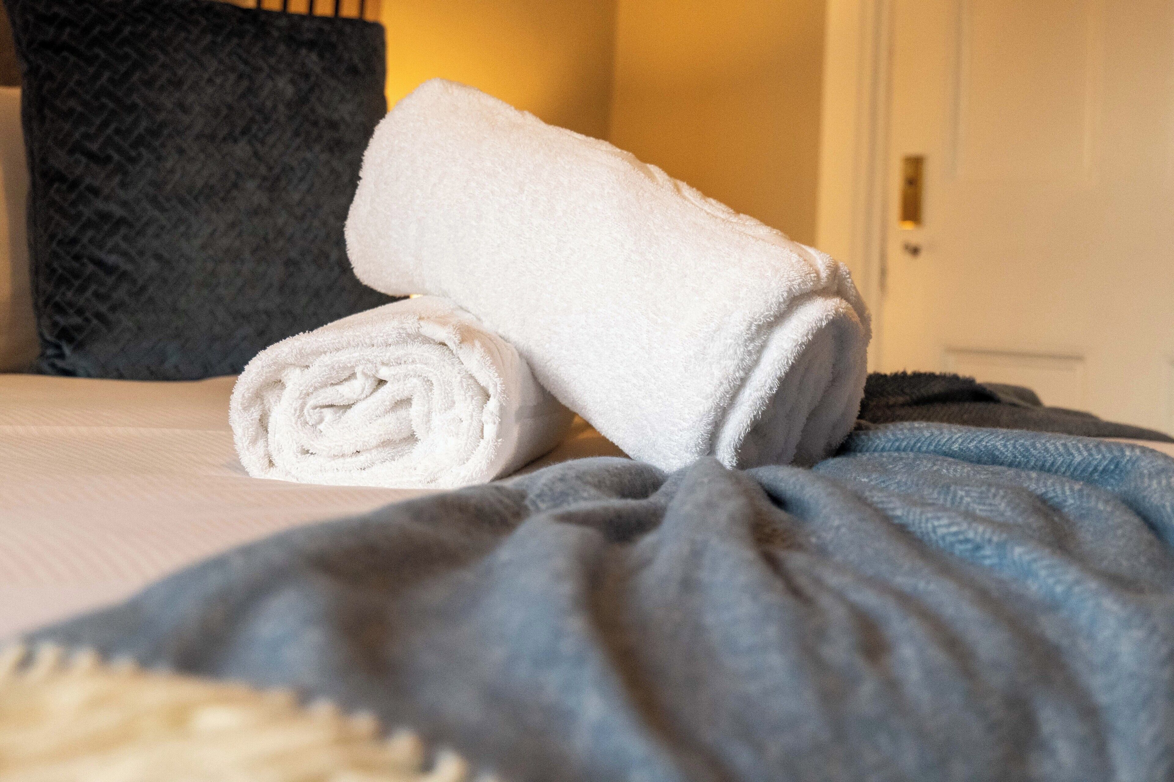 Un lit bien fait, avec une serviette blanche roulée et une serviette blanche pliée, posées sur un lit recouvert d’une couverture grise.