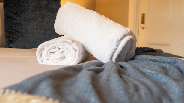 A neatly made bed with a rolled white towel and a folded white towel on a bed with a grey blanket. - The Oliver - Donnini Apartments (Ayr)