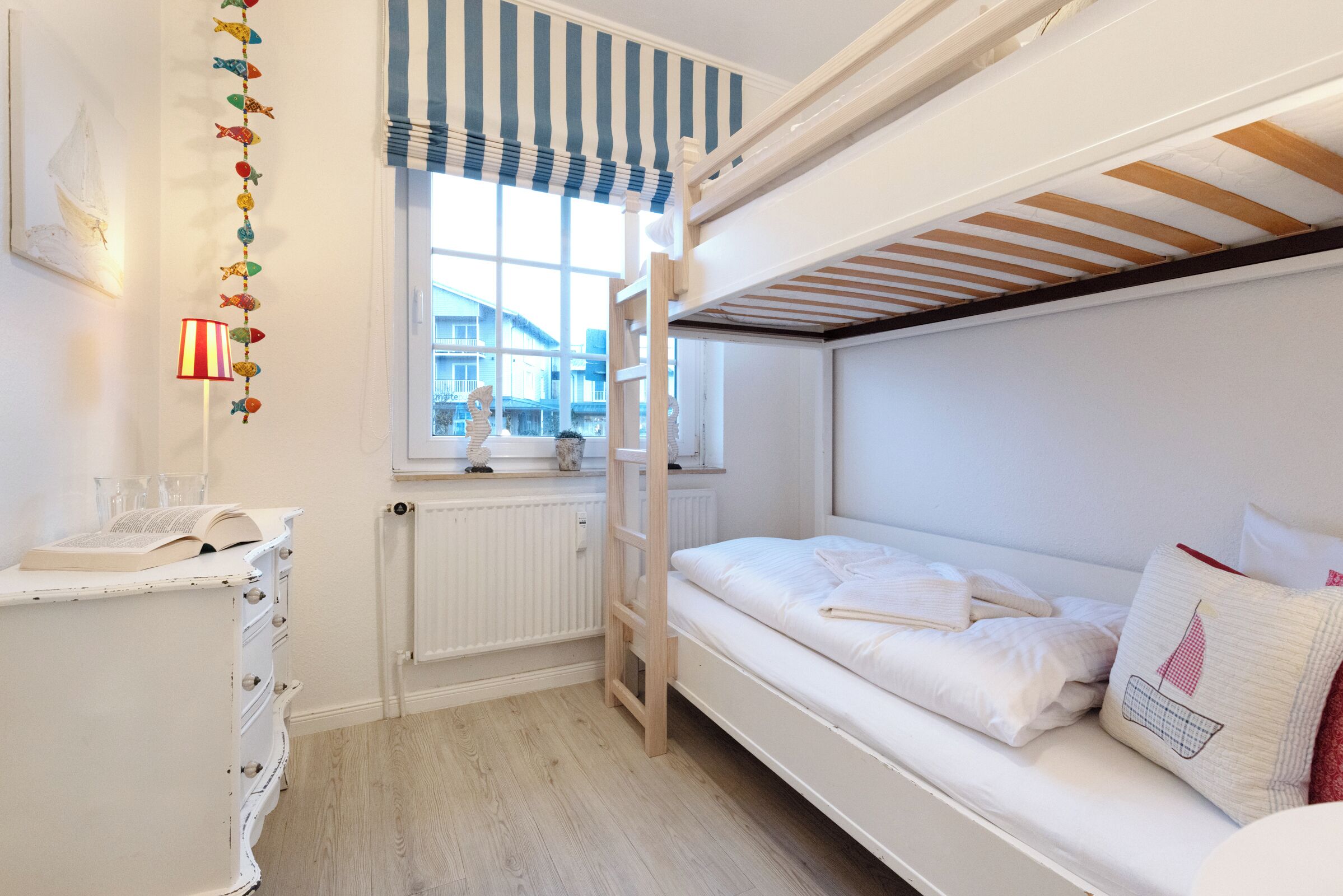 A bunk bed room with a window, a radiator, a dresser with a book, and a decorative fish garland.